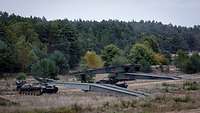 Zwei Panzer stehen nebeneinander und haben vor sich ihre langen Metallbrücke im Gelände abgelegt.