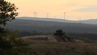 Sonnenaufgang: Ein einzelner Kampfpanzer steht auf einem Hügel, im Hintergrund Windräder.