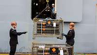 Eine Kiste mit Munition wird unter Aufsicht von uniformierten Personen an Bord eines grauen Schiffes gehoben.