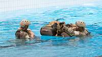 Zwei Soldaten schwimmen in einem Schwimmbecken mit ihrem Diensthund auf einem Rucksackfloss. 