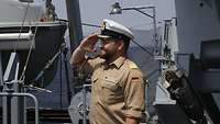 An Bord eines Schiffes steht ein bärtiger Offizier, der salutiert. Er trägt eine markante weiße Mütze mit Goldstickerei.