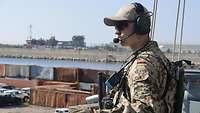 Ein Soldat in Tropentarnuniform, mit Basecap und Headset auf dem Kopf, steht an der Reling und beobachtet die Umgebung.