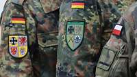 Auf drei Ärmeln von Militäruniformen sind drei unterschiedliche Wappen aufgenäht 