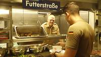 Ein Soldat steht hinter einer Essensausgabetheke. Ein anderer bedient sich am Buffett. Auf einem Schild steht „Futterstraße“.