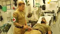 Eine Soldatin mit einer Roten-Kreuz-Armbinde steht neben einer Liege, auf der ein Patient liegt