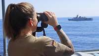 Eine Soldatin steht an der Reling und blickt durch ein Fernglas auf das Meer, auf dem sich ein Schiff befindet