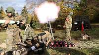 Vier Soldaten schießen auf einer Waldlichtung mit einem Mörser, ein gelber Feuerball entsteht.