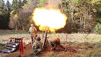 Ein Feuerball steigt aus dem senkrecht stehenden Mörser, an dem sich zwei Soldaten ducken.