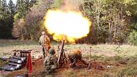 Ein Feuerball steigt aus dem senkrecht stehenden Mörser, an dem sich zwei Soldaten im Kampfanzug vor ein Waldkulisse ducken