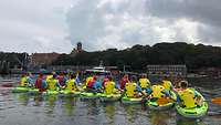 Mehrere Menschen sitzen in Kanus auf dem Wasser. Alle tragen Rettungswesten.