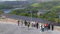 Eine Gruppe Menschen steht auf einer Mauer und auf Häuser und eine Talsperre in der Ferne. 