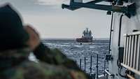 Ein Soldat in Flecktarnuniform auf dem Deck eines Schiffes blickt durch ein Fernglas auf ein rot-weißes Schiff in See.