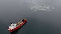 Luftaufnahme einer Wasseroberfläche; ein heller Ring auf einer dunkelblauen Wasseroberfläche; davor ein rot-weißes Schiff.