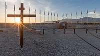Ein Holzkreuz steht im Vordergrund vor vielen Flaggenmasten und einem Gebirge in Afghanistan