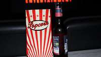 Eine Tüte Popcorn und ein Kaltgetränk stehen auf einer Ablage im Kinosaal, im Hintergrund beginnt der Kinofilm.