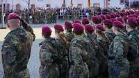 Soldaten sind in Formation auf einem Platz angetreten. Ein Soldat grüßt mit der Hand am Barett. Zuschauer stehen am Rand.