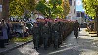 Soldaten marschieren eine Straße entlang. Zuschauer stehen am Rand.