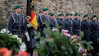 Mehrere Soldaten stehen in Formation vor einer Steinmauer, einer von ihnen hält eine Nationalflagge.