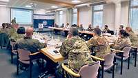 A soldier stands in front of a screen and gives a lecture. Many soldiers sit at tables in a large circle and listen.