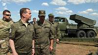 Dmitry Medvedev steht inmitten von Soldaten im Hintergrund ist ein russischer Raketenwerfer zu sehen.