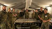 In einem Zelt stehen zehn Soldaten um einen großen Tisch herum und schauen in die Kamera.
