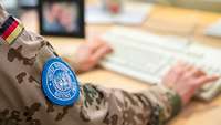 A woman wearing a camouflage-pattern field uniform sits at a desk and taps a keyboard; on her arm, a UN badge can be seen.