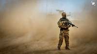 Ein vermummter Soldat mit einem Gewehr steht in einer Staubwolke, im Hintergrund ein Fahrzeug, Dienen in Afghanistan 2020