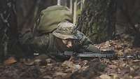 Ein Soldat liegt mit seinem Gewehr und seinem Rucksack auf einem mit Blättern bedeckten Waldboden