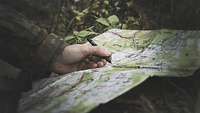 Auf dem mit kleinen Pflanzen bewachsenen Waldboden liegt eine Karte. Ein Soldat zeichnet Markierungen auf der Karte ein.