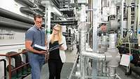 A woman and a man stand next to the technical installations of a building and inspect documents.