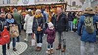 Ein Elternpaar steht mit Ihrer Tochter auf einem Weihnachtsmarkt. 