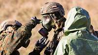 Ein Soldat in Schutzbekleidung beaufsichtigt zwei weitere Soldaten mit ABC-Schutzmaske