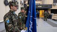 Griechische Soldaten stehen vor dem KFOR-Hauptquatier. Einer der Soldaten hält eine NATO-Flagge in den Händen.