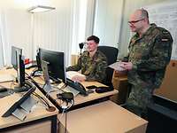 Ein Soldat sitzt an einem Schreibtisch vor einem Computer, neben ihm steht ein anderer Soldat