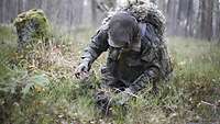 Ein Soldat kniet auf dem Waldboden und tarnt sein Gewehr mit Zweigen und Grashalmen