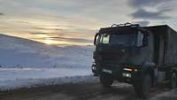 Ein grüner Lastwagen steht auf einer Asphaltstraße in einer verschneiten Landschaft.