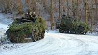 Zwei gepanzerte Kettenfahrzeuge mit darauf sitzenden Soldaten fahren über einen schneebedeckten Waldweg