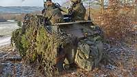 Ein gepanzertes Kettenfahrzeug mit zwei Soldaten und einer Abschussvorrichtung steht an einem Waldrand