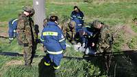 Soldiers in field suits and THW members attach a hose connection. In the background a railway embankment.