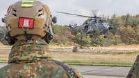 Soldier in field suit with combat helmet stands in front of a low-flying transport helicopter.