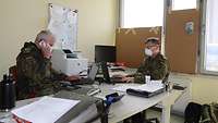 Two soldiers in combat fatigues sit in an office at a table laden with files.