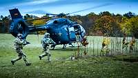 Zwei Soldaten stürmen mit dem Gewehr über eine Waldlichtung. Dahinter landet ein grauer Hubschrauber.