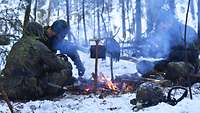 Drei Soldaten sitzen im Wald auf dem Boden, es liegt Schnee. Vor ihnen brennt ein Lagerfeuer, an dem sie sich wärmen.