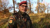 Ein Soldat im Feldanzug mit rotem Barrett steht vertrauensvoll lächelnd im Wald und blickt in die Kamera