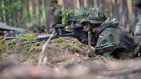 Zwei Soldaten liegen nebeneinander mit der Waffe im Anschlag im Wald in einer Stellung.