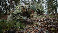 Zwei Soldaten knien am Waldboden und bearbeiten ihre Tarnkleidung