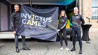 Eine Frau und ein Mann halten eine Flagge mit dem Logo der Invictus Games Düsseldorf 2023 hoch. Ein anderer Mann steht daneben.