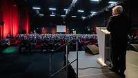 Ein Soldat steht am Rednerpult mit Blick auf ein sehr großes Auditorium. 