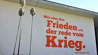 Auf einer Wand steht der Satz „Wer aber den Frieden will, der rede vom Krieg“ in roter Schrift.