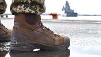 Stiefel eines Soldaten spiegeln sich im Vordergrund in einer Pfütze. Im Hintergrund steht ein Kriegsschiff am Hafen.